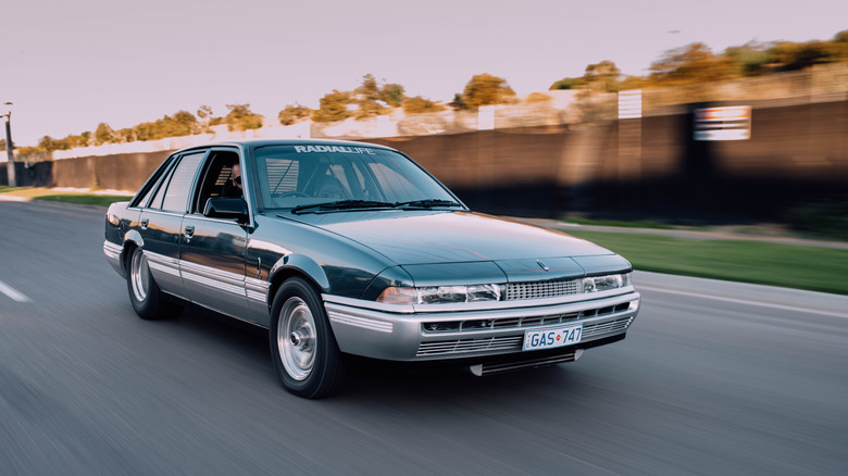 A black and silver Holden Calais VL driving on a road past a fence and grass