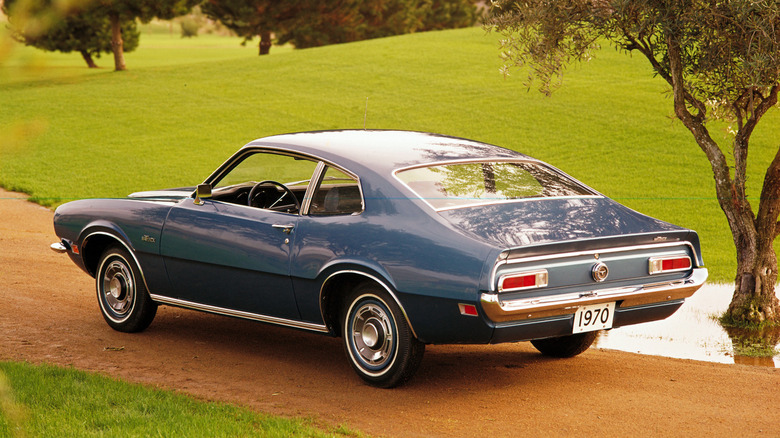 A catalogue photo of Ford's 1970 Maverick, blue with chrome accents on trim, edges and bumper, sitting on a dirt roadway surrounded by well-manicured greenery.