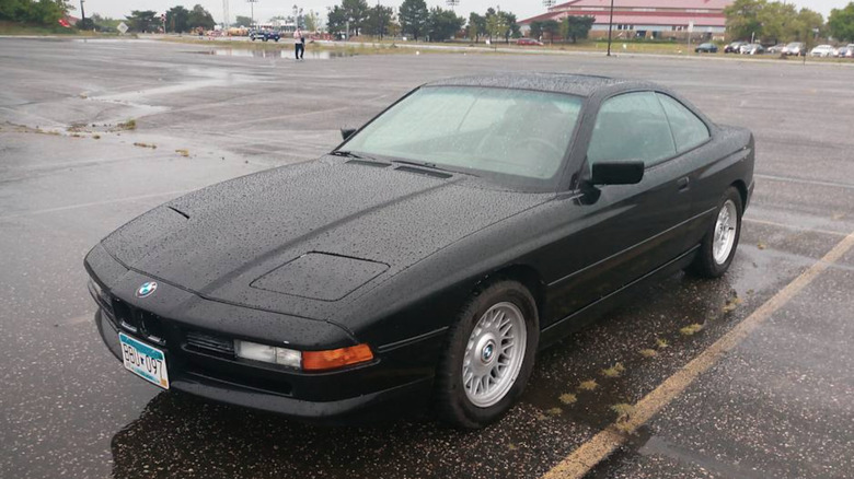 A gorgeous black BMW E31 dusted in rain drops, sitting on a wet parking lot tarmac