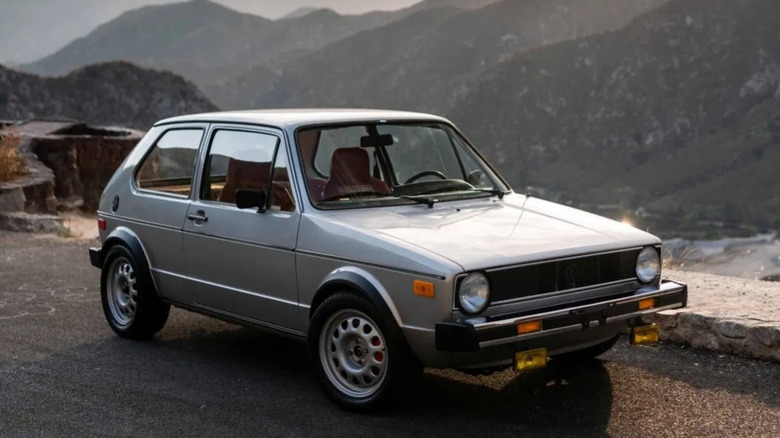 A silver, two-door 1978 Volkswagen Rabbit on a paved road with mountains in the background.