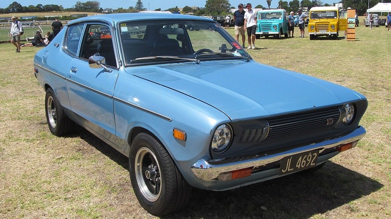 A powder blue 1978 Datsun B210 sitting on some dead grass at a car event.