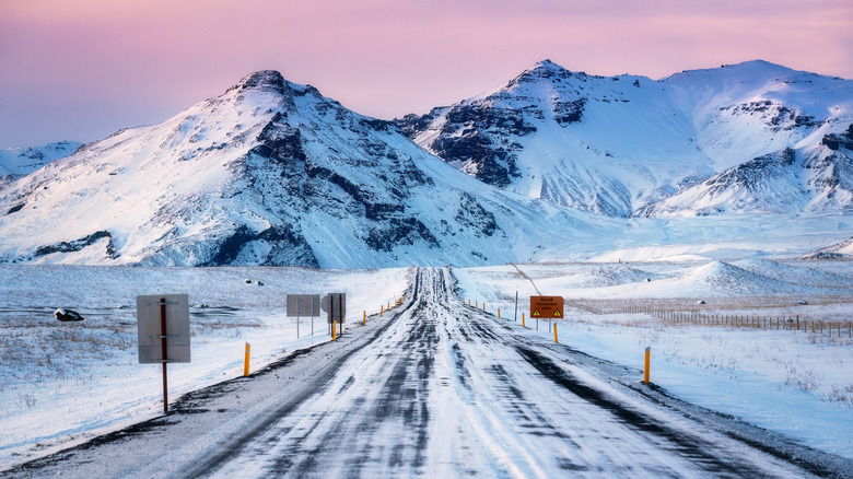 Scenic view from Ring Road Route 1 in Iceland during winter at sunset, with pink clouds, road signs, poles on the side of road, mountains in the background, driving through the beautiful Iceland