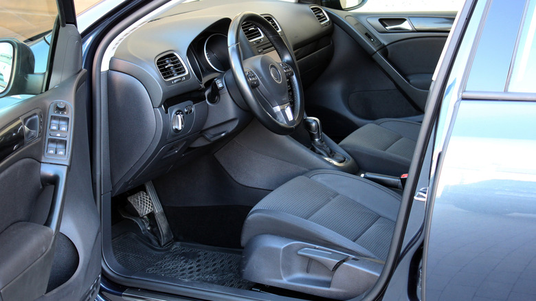Modern car interior with open drivers door, featuring black fabric seats, leather-wrapped steering wheel, textured plastic dashboard, and chrome trim around controls and handles.