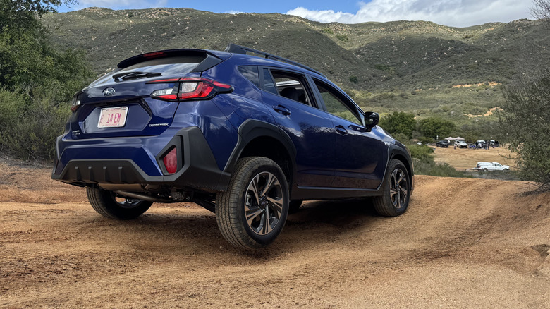 Rear three-quarters shot of a blue Subaru Crosstrek stopped on a dirt trail with its rear wheel cocked up and green mountains in the background