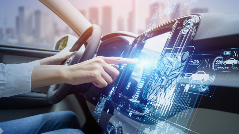 A woman's finger comes across the frame to touch a vehicle touchscreen with different data illustrations in blue lighting surrounding the touch screen on the dashboard.