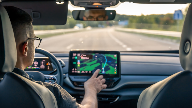 Man touches a car touchscreen from the driver's seat. The screen is a map from a navigation application.