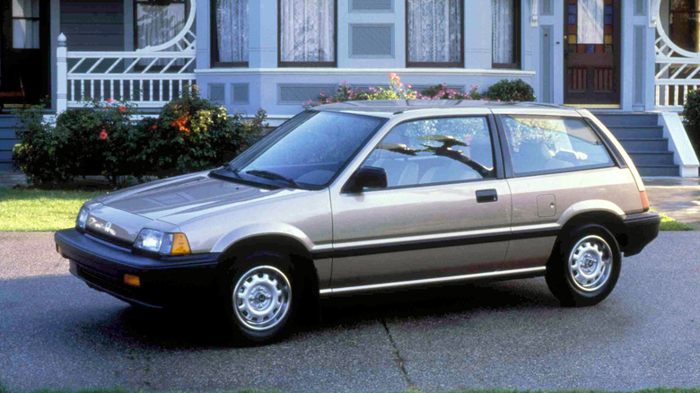 A gold third generation Honda Civic Hatchback, 1984, sitting in a driveway in front of an older victorian style home.