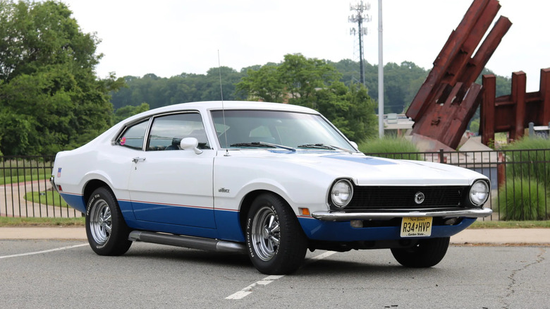 A front three-quarters shot of a white '70 Ford Maverick parked in a lot in front of a fence, trees, and a metal structure