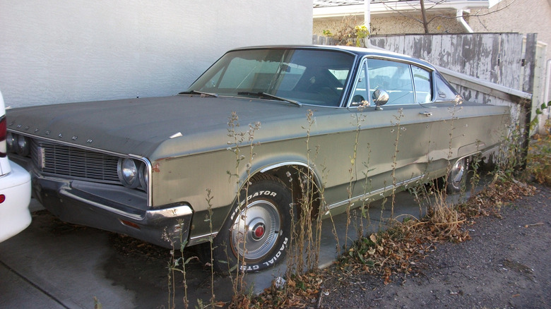 A front three quarters shot of a run-down '68 Chrysler Newport Custom Coupe parked next to a white building with weeds growing next to it