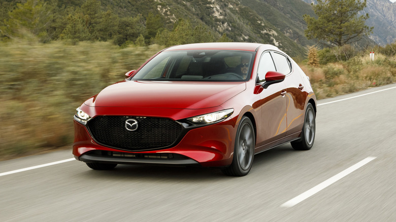 A front three-quarters shot of a red Mazda3 hatchback driving on a mountain road past trees and shrubs