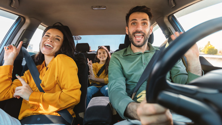 An overenthusiastic dad sings/dances with his wife and child in the car.