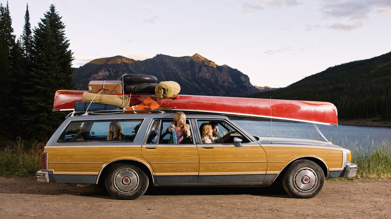 A woody station wagon with a family sitting inside, canoe and luggage affixed to the top of the car, sitting beside a lake.