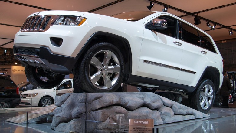 A white 2011 Jeep Grand Cherokee parked on a fake rock formation at a dealership.