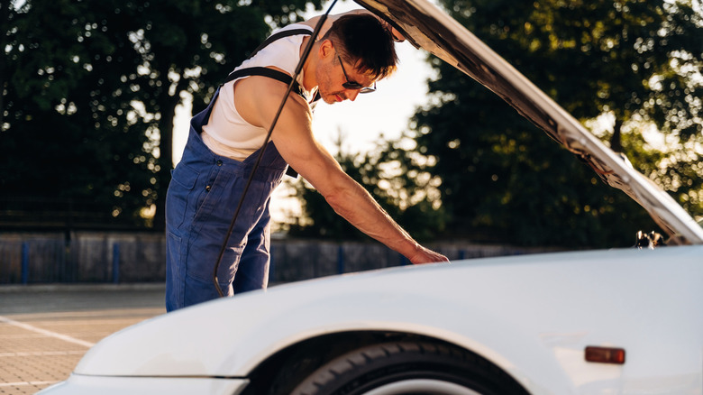 Male mechanic checks engine components of a car in an outdoor parking area, with a raised hood and trees visible in the background  by: Standret
