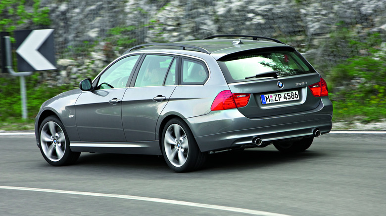Rear three-quarters shot of a siler BMW 335i wagon driving on a road past a wall of rock and a black and white arrow sign