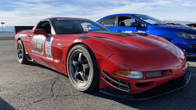 Front three-quarters shot of a modified red C5 Corvette parked on asphalt in front of a blue Subar WRX and a cloudy sky