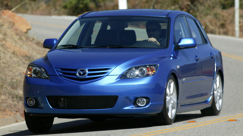 Front three-quarters shot of a blue '04 Mazda3 hatchback driving around a corner on a mountain road past brown mountainsides
