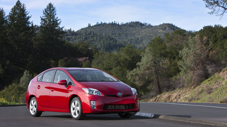 A red 2010 Toyota Prius parks on pavement among wooded hills.