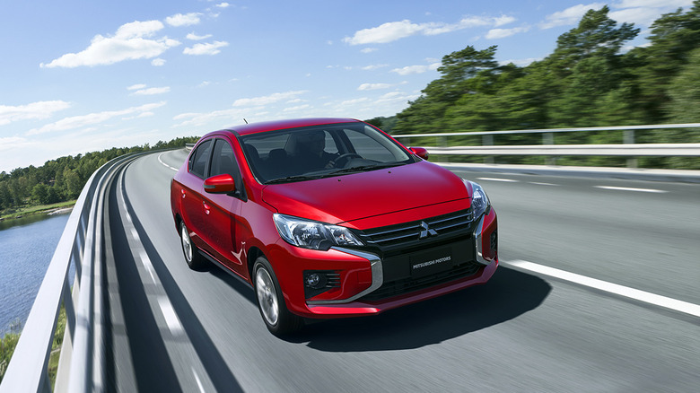 A red Mitsubishi Mirage rounds a bend over a body of water.