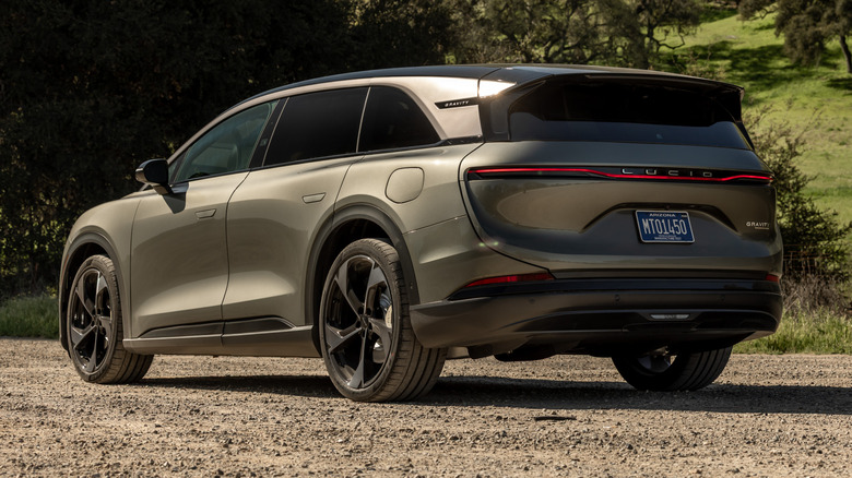 A rear three-quarter view of a bronze Lucid Gravity EV parked on a gravel surface with greenery in the background.