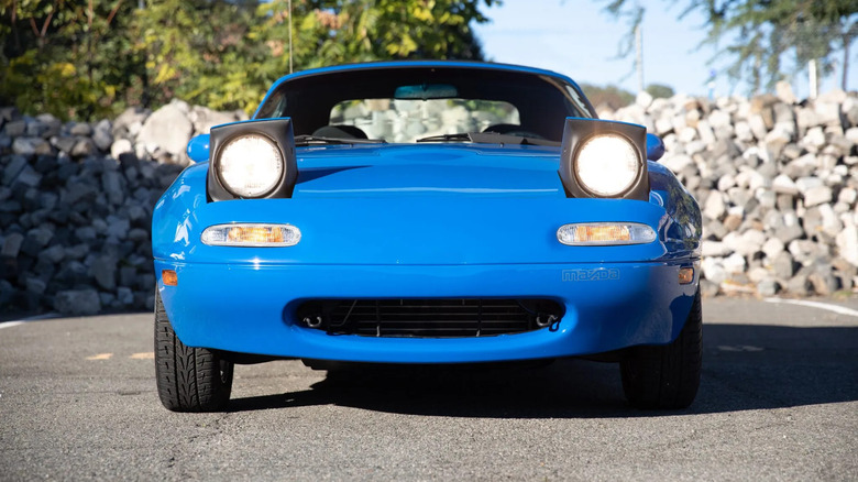 A head-on, close-up shot of a blue NA Miata with its headlights popped up