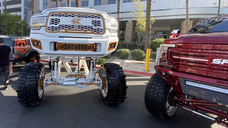 A photo of two brodozers, one white and gold Ram HD that's lifted like 8 ft off the ground and a red Ford Super Duty