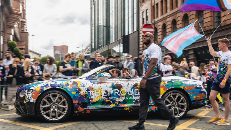 Side view of a Bentley Continental GTC with rainbow graphics driving in a parade with a man walking next to it and the Trans Pride flag