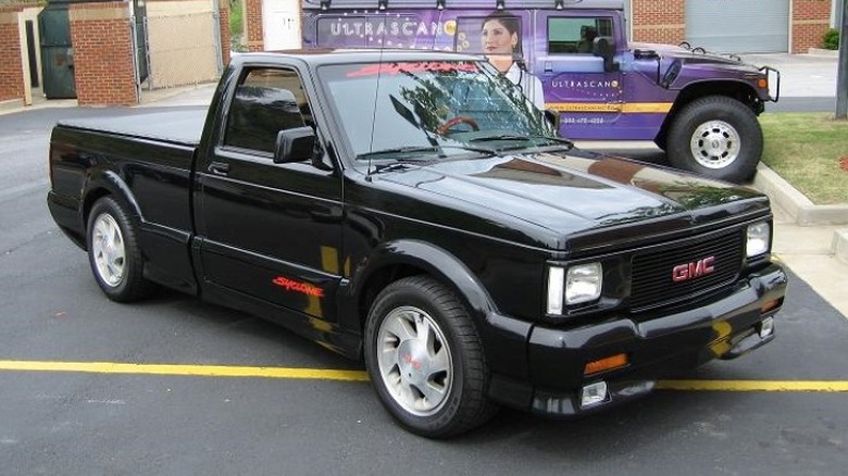 A black 1991 GMC Syclone parked in an outdoor parking lot with a purple Hummer in the background
