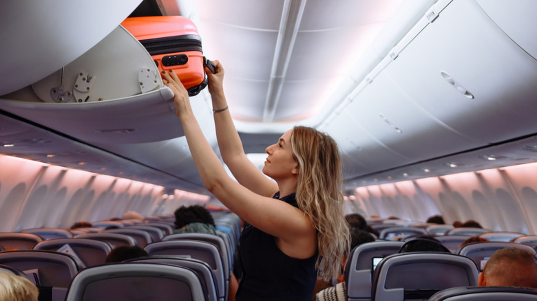 woman puts an orange bag into overhead bins on an airplane