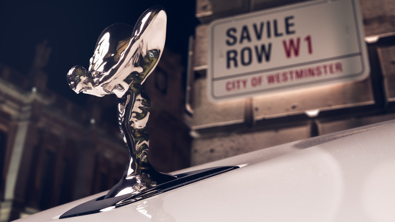 A close up of the Spirit of Ecstasy on the hood of a white Rolls in front of a Savile Row sign