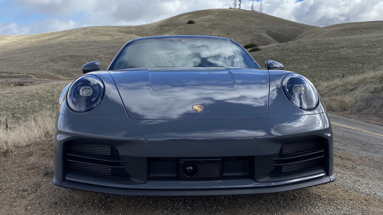 A head-on shot of a dark gray Porsche 911 showing its round headlights in front of rolling hills covered in dead grass