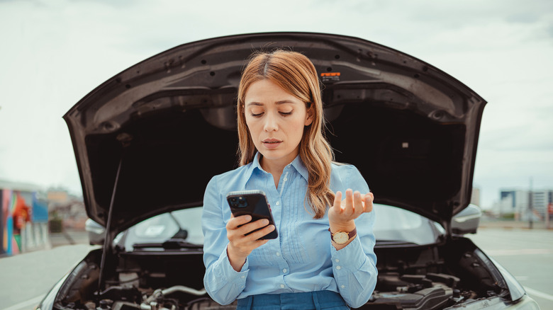 Woman leaning up against hood of car with open engine bay, making a call on her phone.
