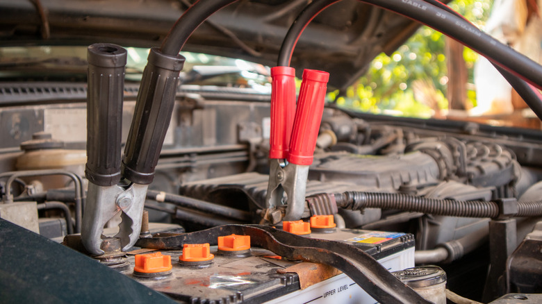 A pair of clamps, one with red handles, one with black handles, are attached to a car battery, housed in a vehicle's engine bay.