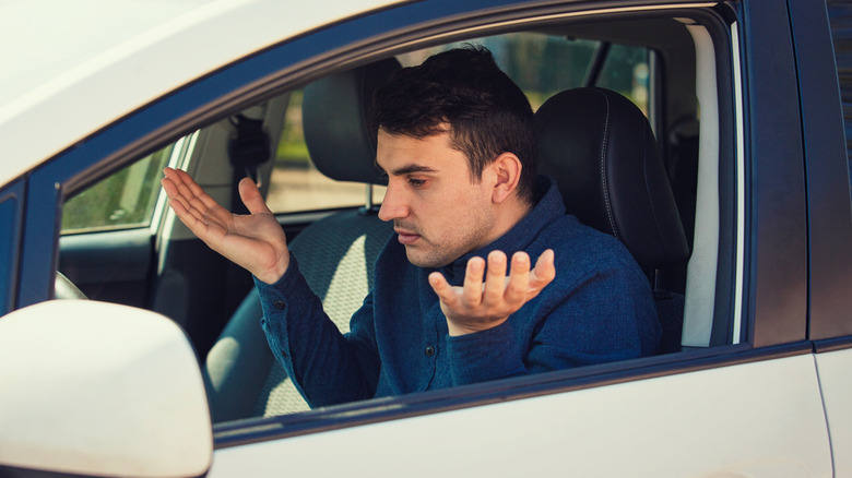 Angry young man driver shaking hands and shrugging shoulders, has problems with the car. Displeased perplexed guy has a road accident. Traffic jam rush hour.