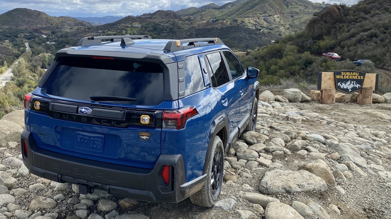 A rear three-quarters shot of a blue 2026 Subaru Outback Wilderness parked on rocks in front of a view of more mountains and a Subaru Wilderness sign