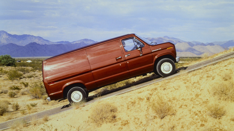 A red 1982 Ford Econoline driving up a hill with mountains in the background
