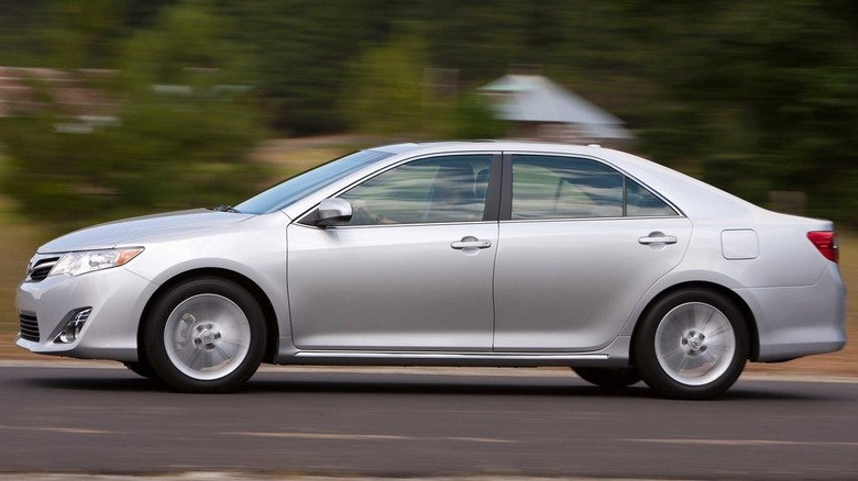 A side view of a silver Camry driving past trees and a home