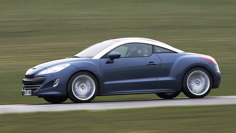 A side view of a dark blue and silver Peugeot RCZ driving on a road past a blurry grassy field