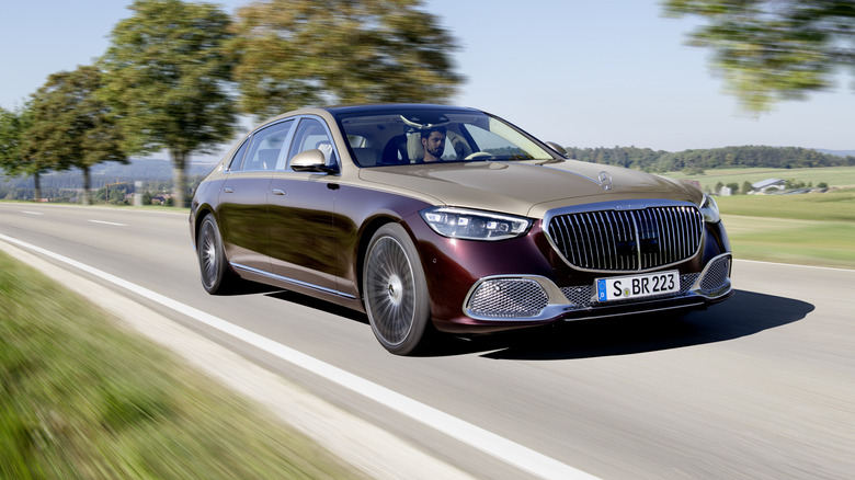 A front three quarters shot of a gold and burgundy Maybach S-Class driving on a rural road past trees and grassy fields