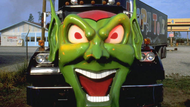 A green demon-like mask sits over the grill of a black semi that appears to be transporting "toyz" according to the font on the side of the truck, parked at a derelict gas station. A still from the film Maximum Overdrive.
