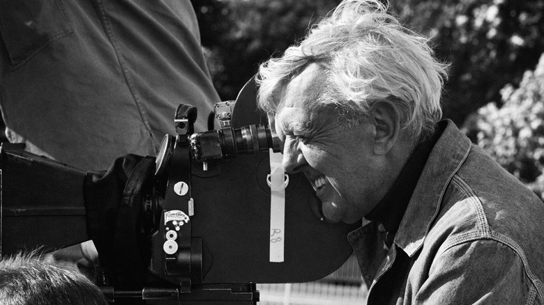 Jaques Tati looks through the viewfinder of a motion picture camera, in black and white.
