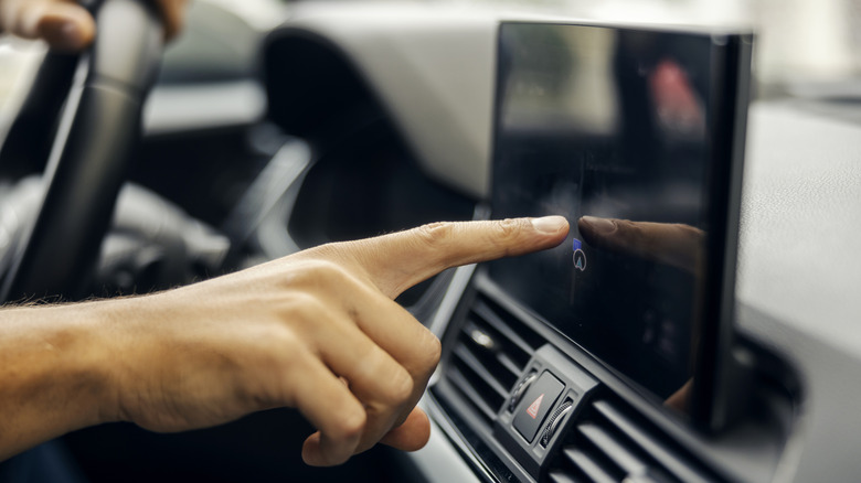 Person's hand interacting with a modern in-car touchscreen display, selecting an option on the control panel for navigation and infotainment features while driving