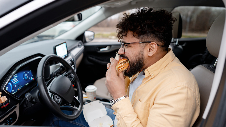 Young adult man enjoying takeout burger in driver's seat of modern car