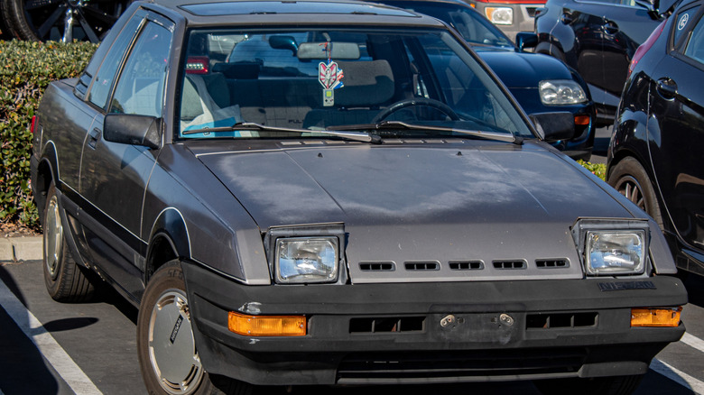front view of a sun damaged gray Nissan Pulsar NX with its headlights popped up in a parking lot in front of a hedge