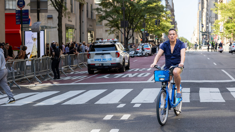 Man on a rental bicycle with a half-opened shirt and shorts cycles along a busy avenue closed to traffic. A police car is on the side.