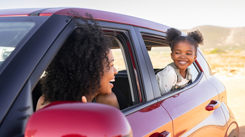Cheerful little african american daughter sitting on back seat of car looking at mother from mirror while travelling. Young fun woman with cheerful female kid enjoying road trip. Traveling in car.