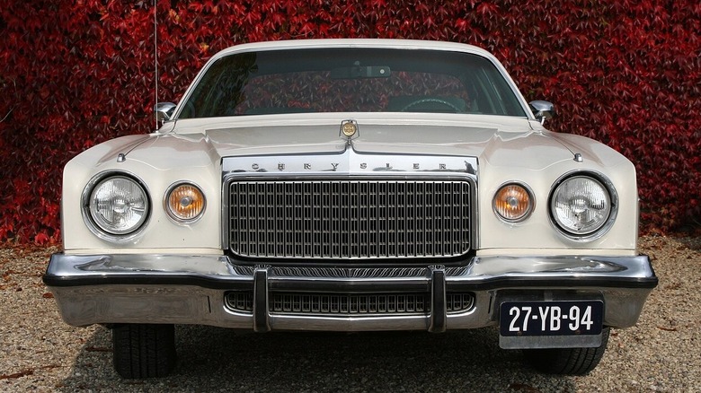 The front end of a white Chrysler Cordoba with a wall of red ivy plants behind.