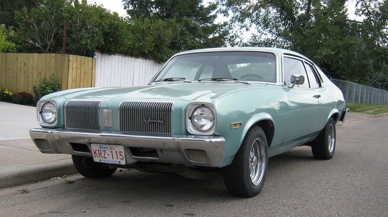 A light teal blue Oldsmobile Omega parked on a neighborhood street with  wood fencing behind it.