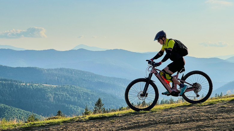 Cyclist man riding electric mountain bike outdoors. Male tourist biking along grassy trail in the mountains, wearing helmet and backpack. Concept of sport, active leisure and nature.