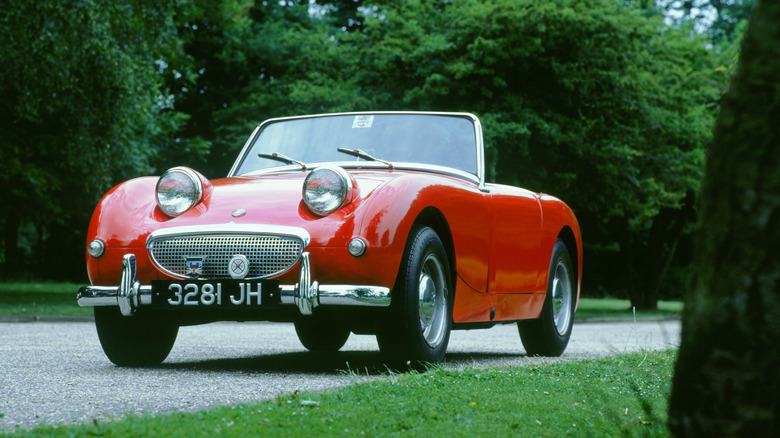 A red austin healey sprite parked in a driveway surrounded by green leafy trees